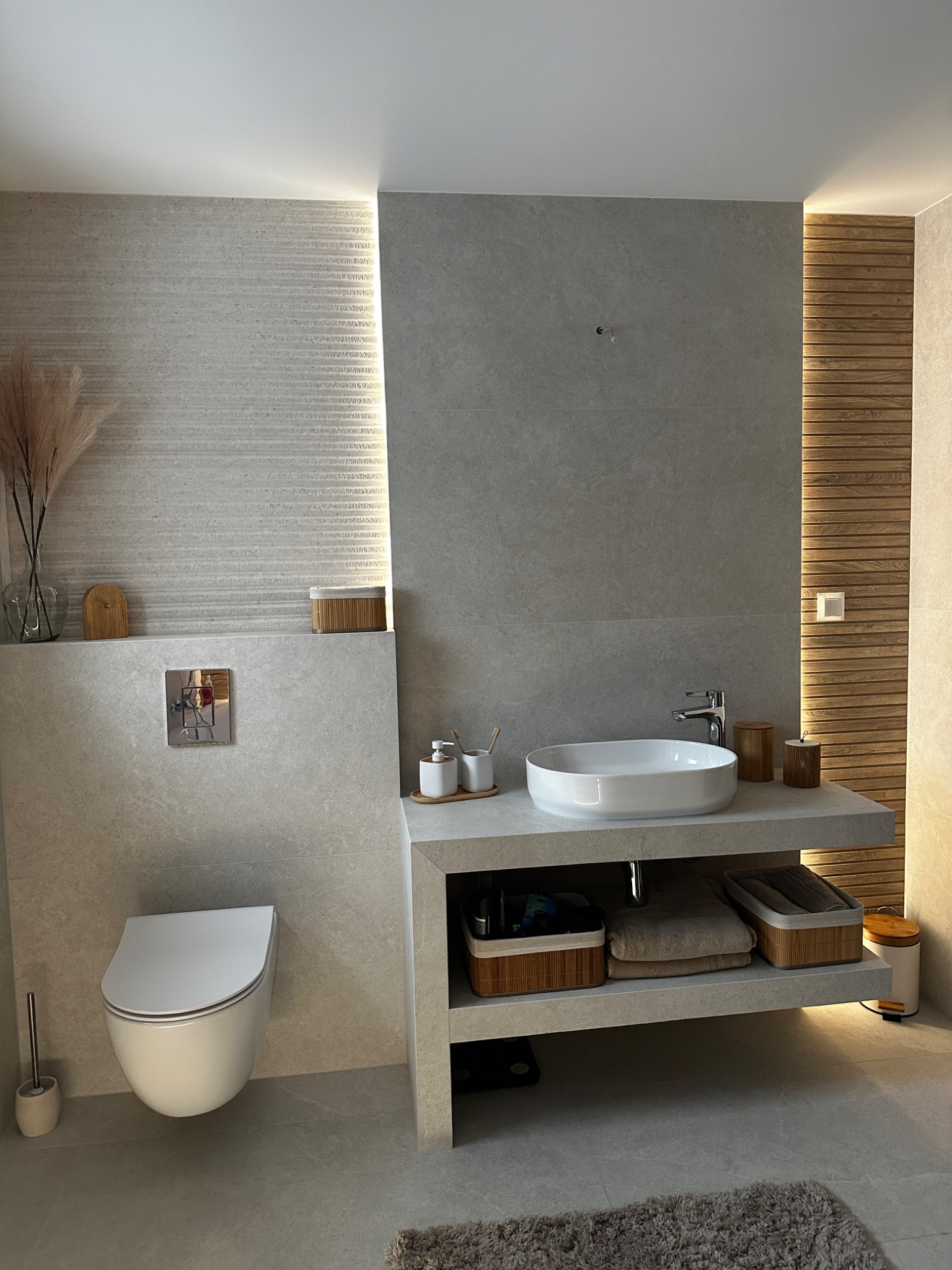 Nowoczesna łazienka z wiszącą toaletą, umywalką nablatową i podświetlanymi panelami drewnianymi, wykończona w odcieniach szarości i beżu, z dekoracyjnymi trawami pampasowymi w wazonie.