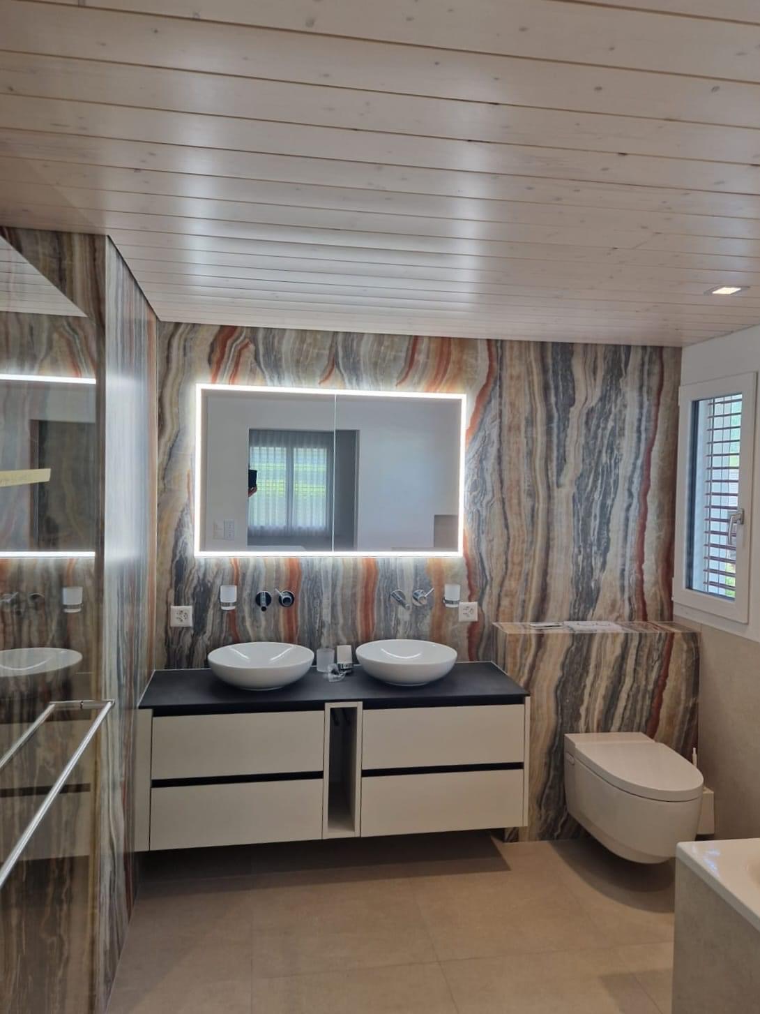 Nowoczesna łazienka z podwójną umywalką nablatową, lustrem z podświetleniem LED, oraz ścianami wykończonymi płytkami imitującymi marmur w odcieniach szarości, brązu i czerwieni. Sufit wykończony...