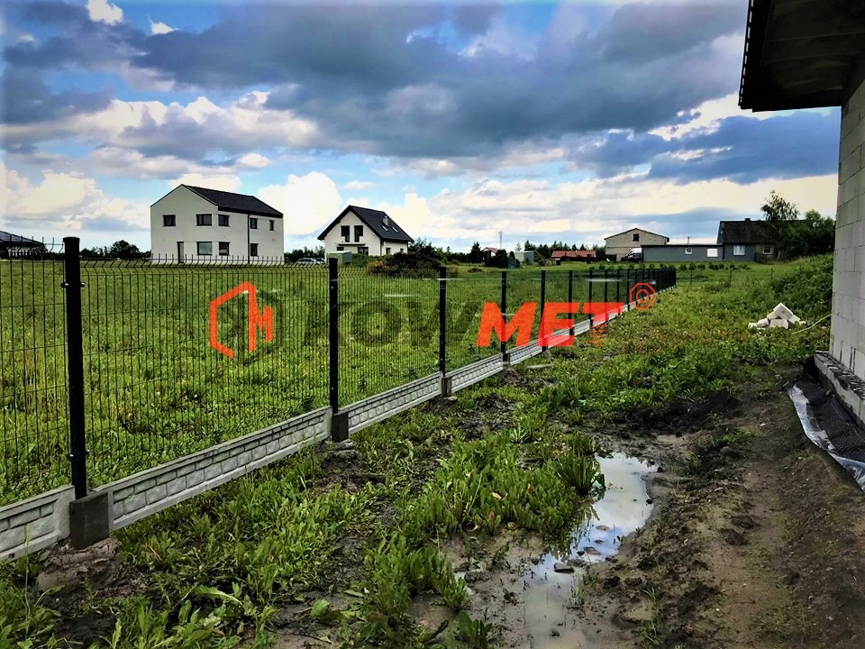 Nowo postawione ogrodzenie z siatki z podmurówką betonową na nierównym terenie po deszczu, w tle domy jednorodzinne i pochmurne niebo.