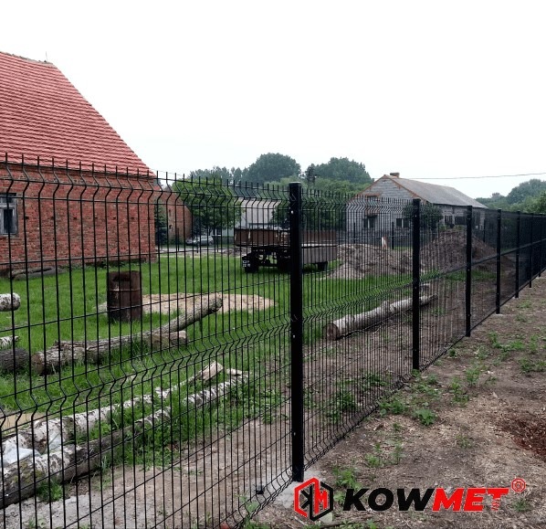 Czarne, metalowe ogrodzenie z paneli 3D otacza posesję z widocznymi budynkami gospodarczymi i starym domem z czerwonej cegły. Na trawniku leżą pocięte pnie drzew.