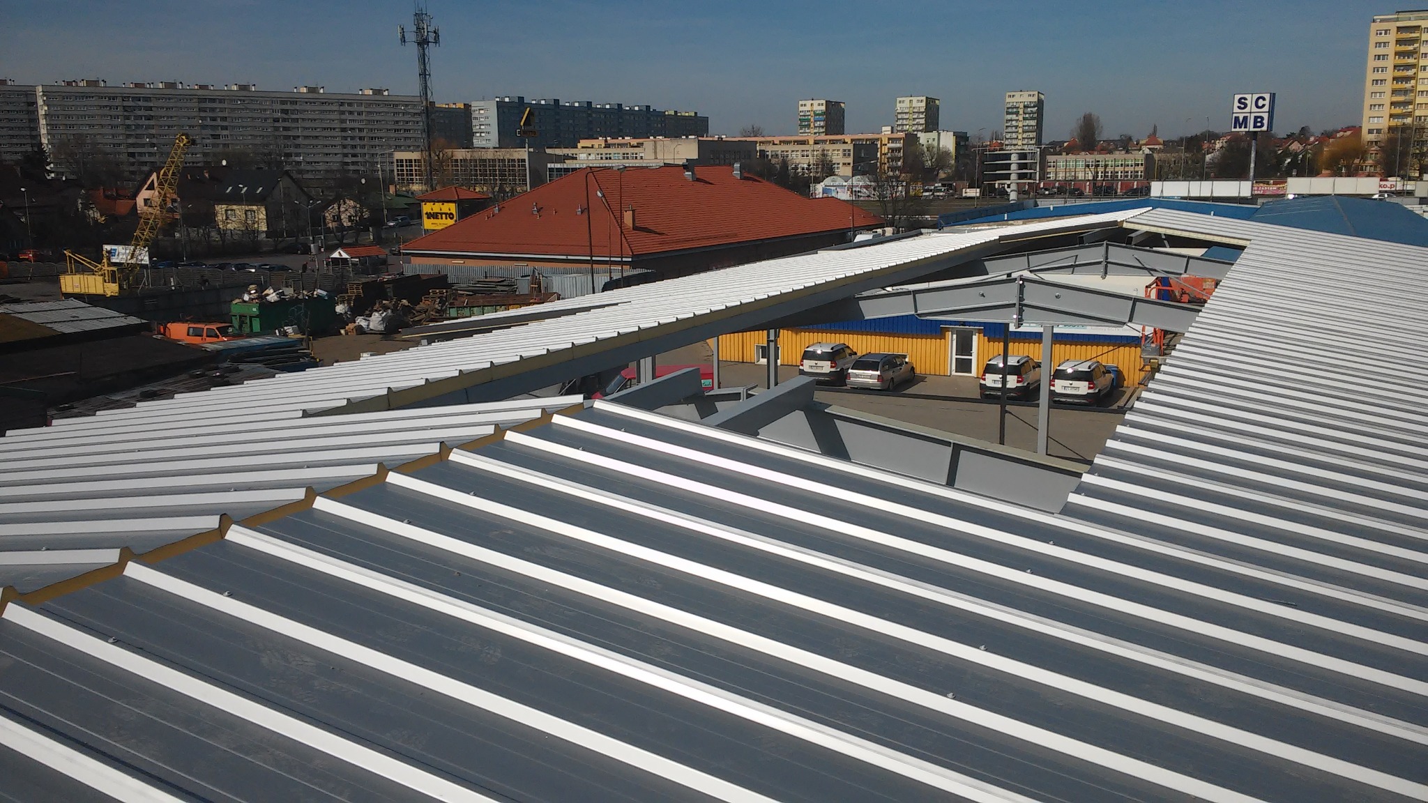 Nowe, szare blaszane pokrycie dachowe z białymi pasami, widok z góry na tle zabudowy miejskiej i błękitnego nieba.