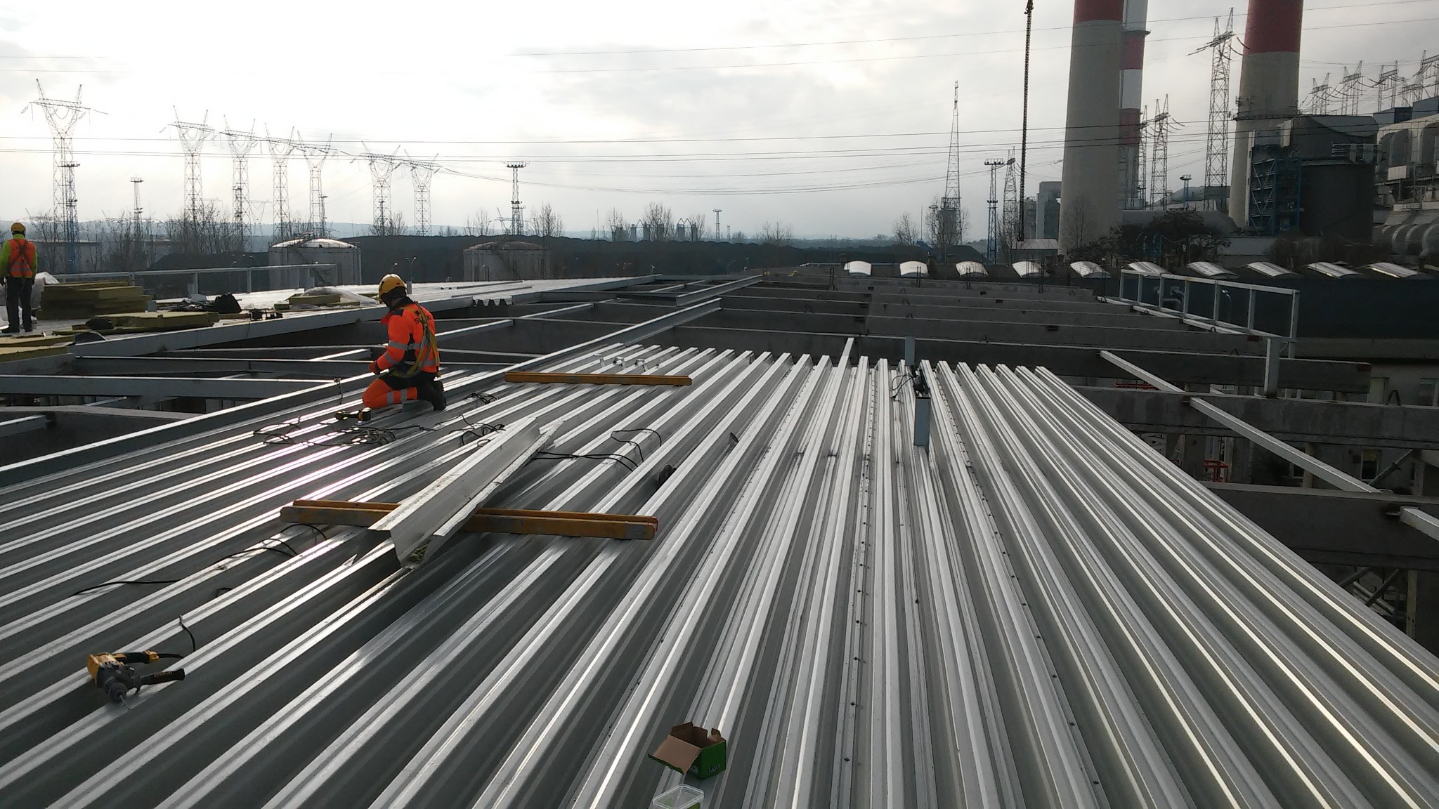 Pracownik w pomarańczowym kombinezonie klęczy na metalowej konstrukcji dachu z widocznym narzędziem, w tle kominy przemysłowe i słupy wysokiego napięcia.