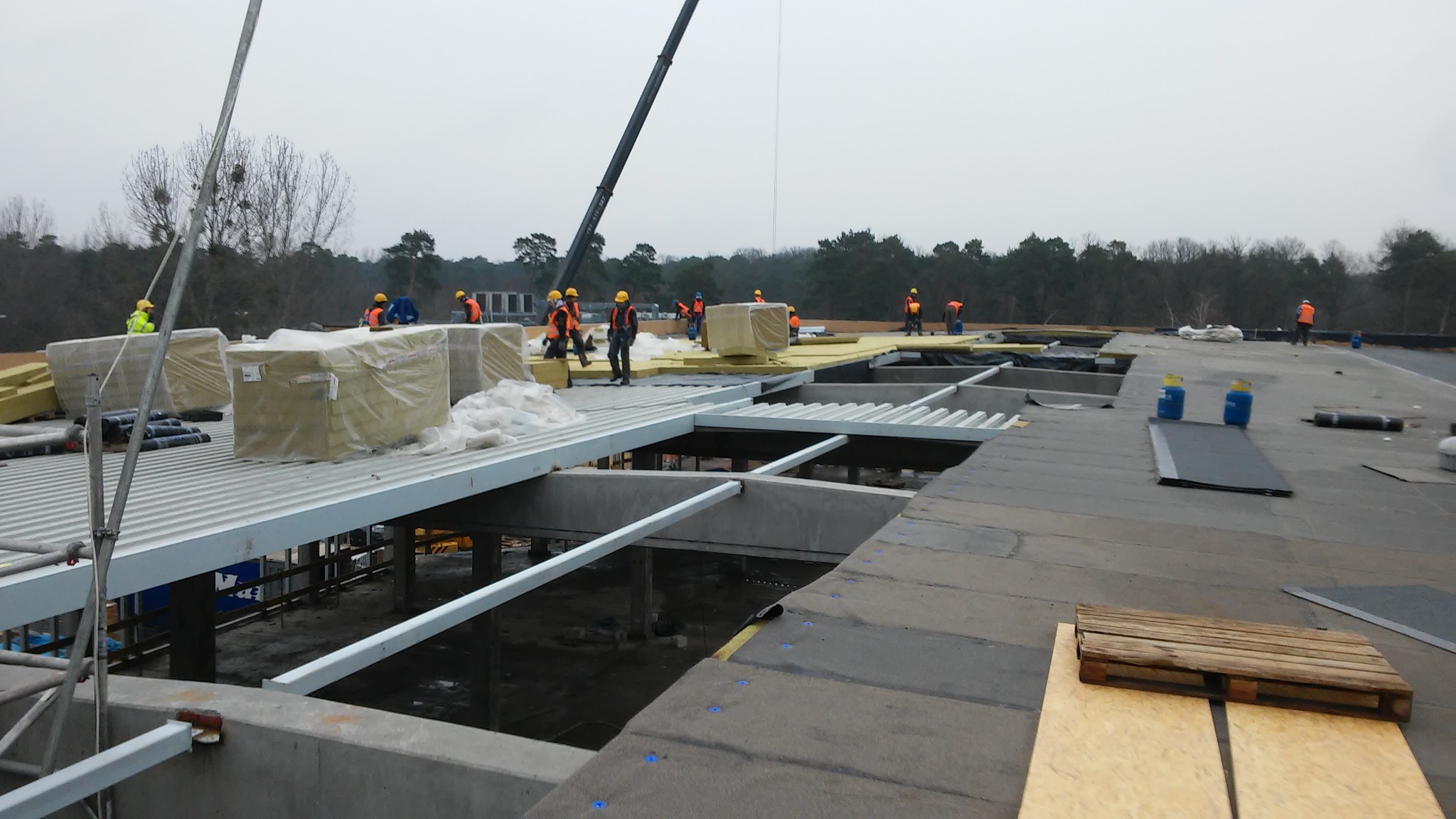 Montaż izolacji termicznej na dachu przemysłowym z widocznymi pracownikami w kamizelkach odblaskowych, dźwigiem oraz materiałami izolacyjnymi w folii ochronnej.