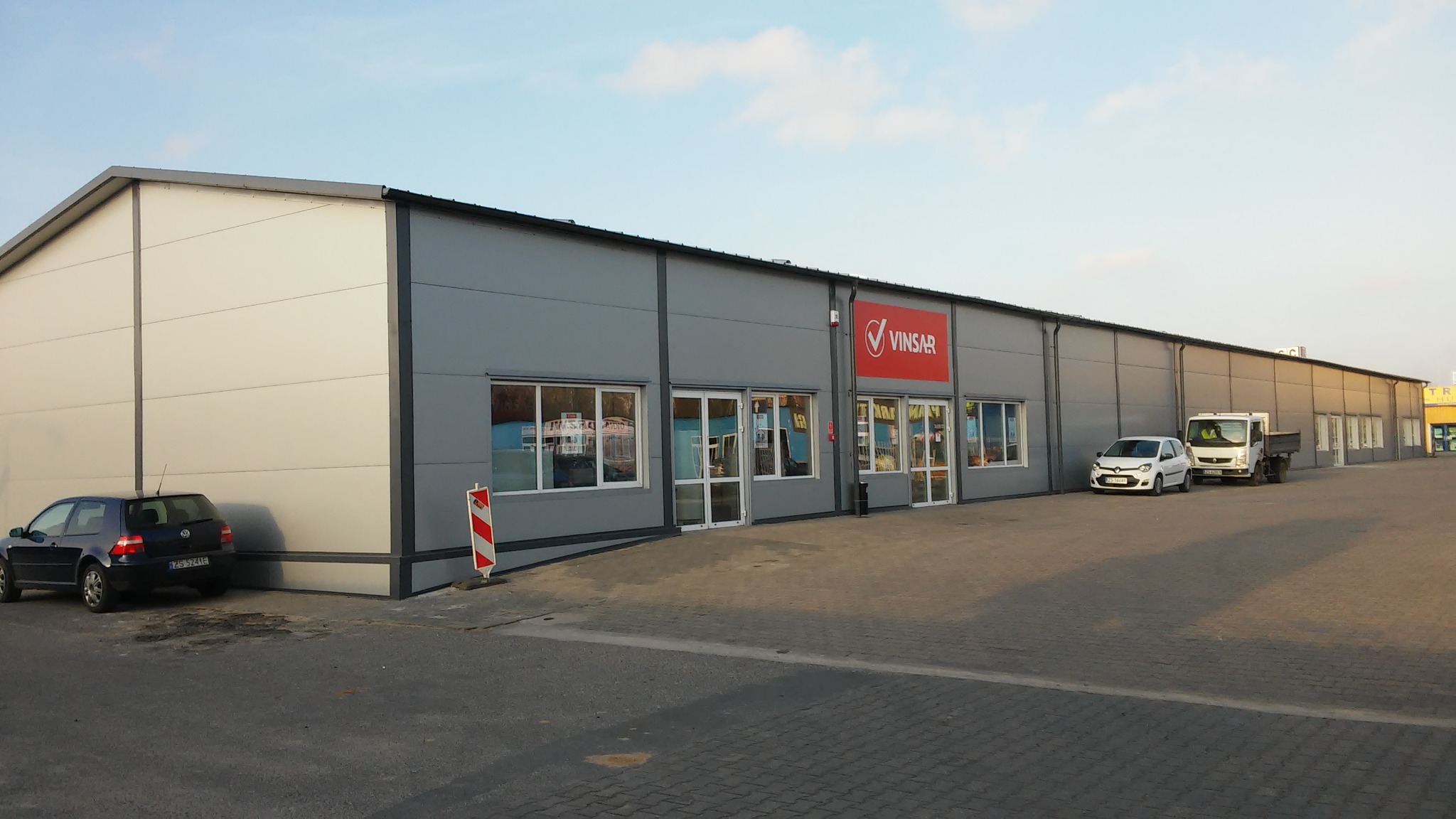 Szary budynek magazynowy z logo firmy Vinsar nad wejściem, widoczne samochody osobowe i dostawcze na parkingu.
