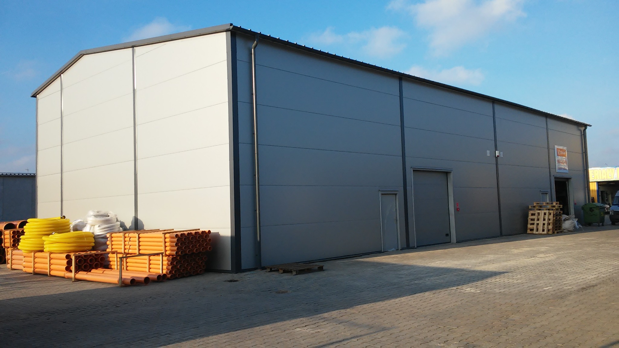 Nowoczesny magazyn z paneli warstwowych w kolorze szarym i białym, z widocznymi rynnami i stosem pomarańczowych rur PCV na zewnątrz, w słoneczny dzień.