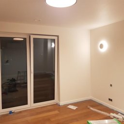 Paulina Skalny Skalbud - Pomalowane na biało wnętrze pokoju z drewnianą podłogą, widoczne przesuwne drzwi balkonowe, okrągła lampa ścienna i listwy przypodłogowe zabezpieczone niebieską taśmą malarską.