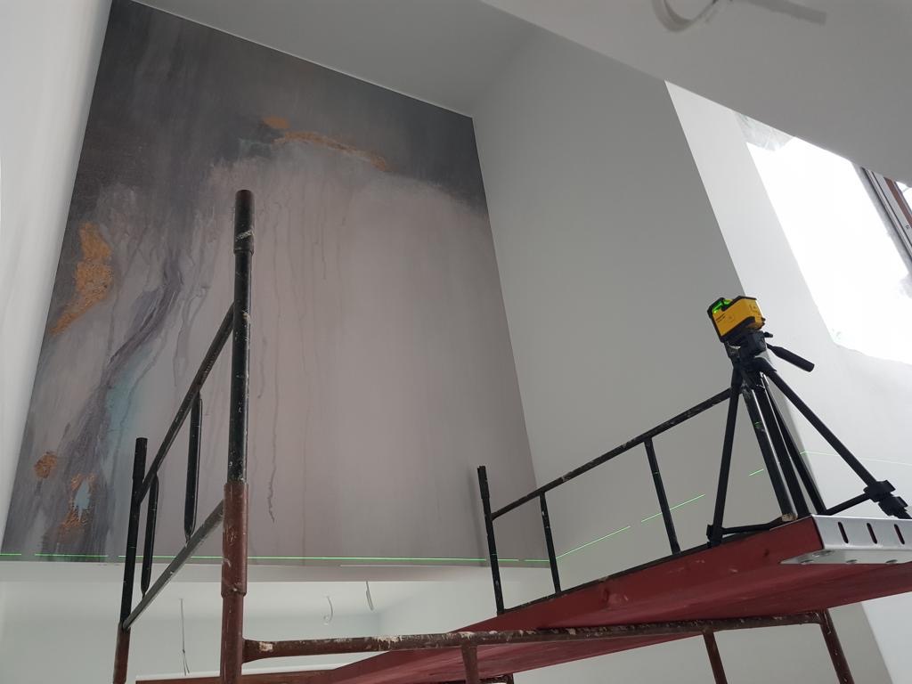 Ściana z abstrakcyjnym wzorem w odcieniach szarości i złota, oświetlona laserowym niwelatorem ustawionym na rusztowaniu, widoczne zielone linie lasera.