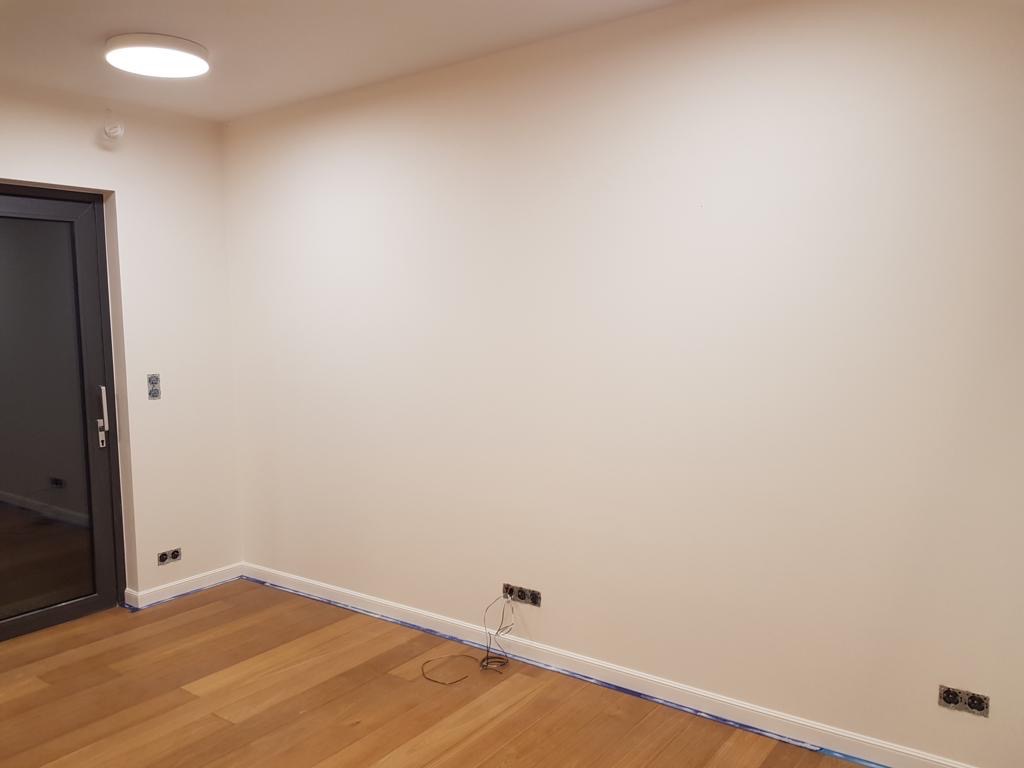 Pokój po świeżym malowaniu ścian na jasny odcień, widoczne listwy przypodłogowe zabezpieczone taśmą malarską, drewniana podłoga i gniazdka elektryczne.