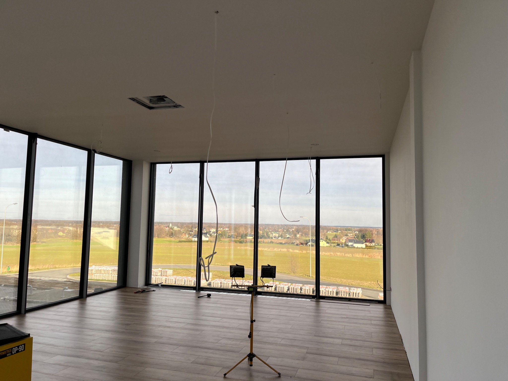Wnętrze biura z dużymi oknami, drewnopodobną podłogą i lampami budowlanymi na statywie, widoczne przygotowania do malowania ścian.