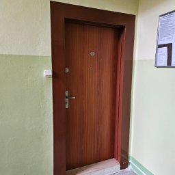 Krystian Kuna - Brązowe, drewnopodobne drzwi wejściowe z numerem '50' w brązowej futrynie, na tle ściany w odcieniach zieleni i beżu, w korytarzu z szarym lastriko.