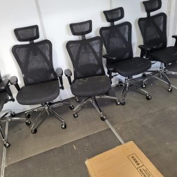 Krystian Kuna - Rząd czarnych, siatkowych krzeseł biurowych z zagłówkami i chromowanymi podstawami na kółkach, ustawionych na szarej podłodze, z kartonem w rogu.