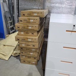 Krystian Kuna - Stos kartonów z oznaczeniami 'GLAS!BREAKABLE!FRAGILE!' obok białej komody z szufladami w surowym wnętrzu. Widoczna drabina i folia izolacyjna na ścianie.