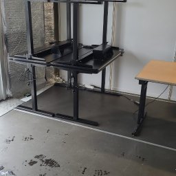 Krystian Kuna - Montaż czarnego, regulowanego biurka z metalową ramą. Widoczne kable i elementy konstrukcyjne. Podłoga z plamami i śladami. Częściowo zmontowany mebel.