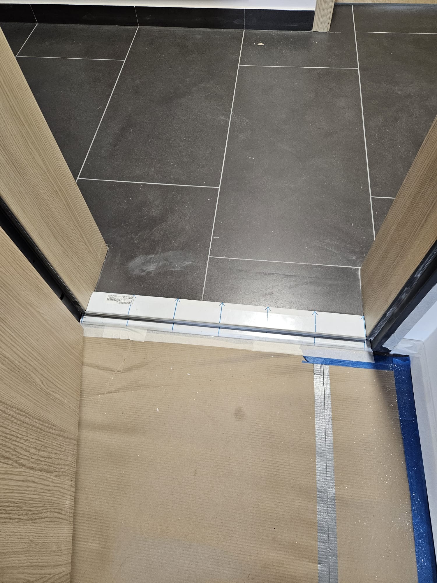 Szare płytki podłogowe ułożone w prostej jodełce, widoczne przez otwarte drzwi z drewnianą okleiną i metalową listwą progową, zabezpieczone kartonem i niebieską folią.