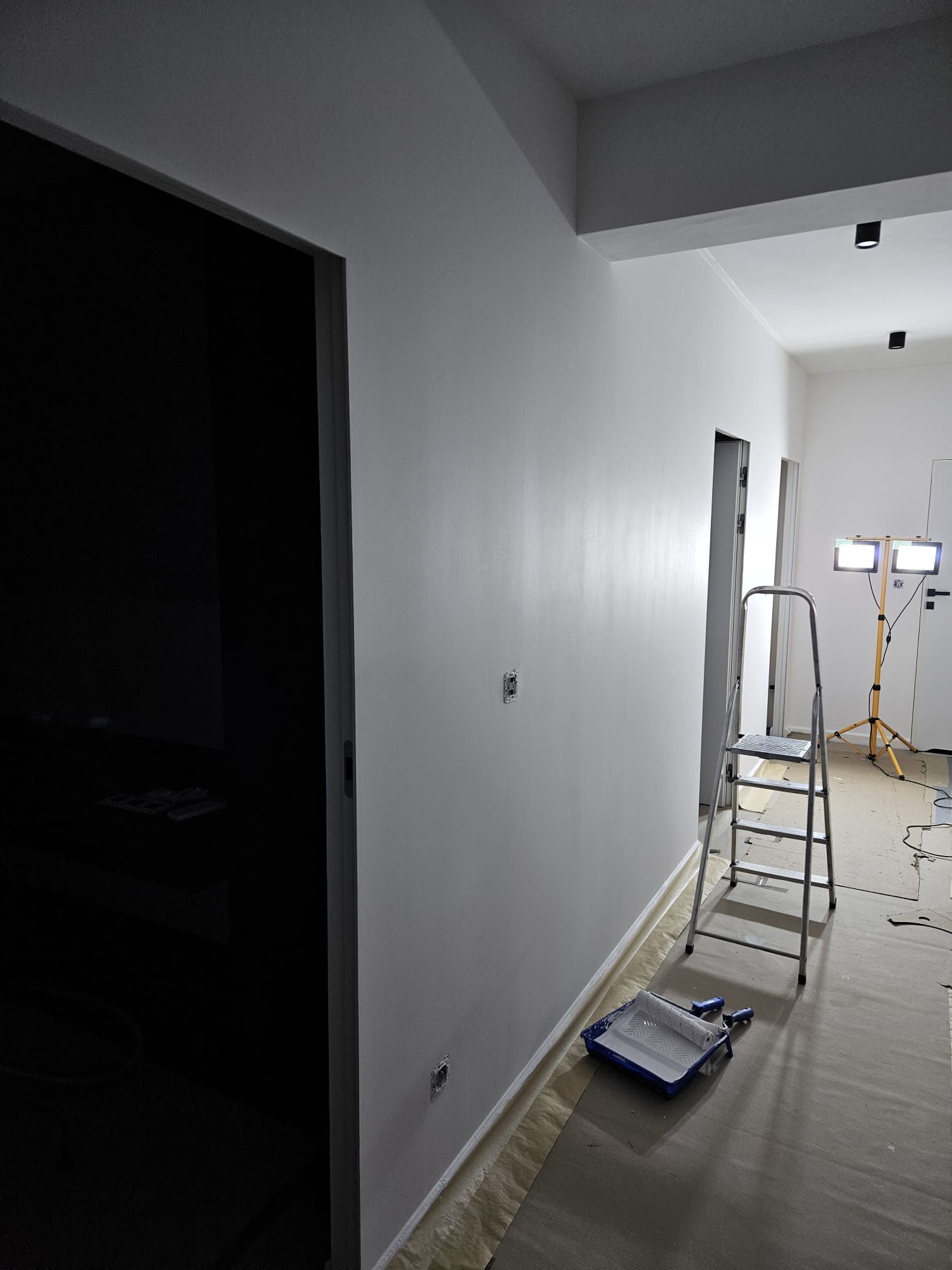Świeżo pomalowana na biało ściana w korytarzu, drabina, kuweta z wałkiem malarskim i lampy halogenowe oświetlają wnętrze podczas prac wykończeniowych.