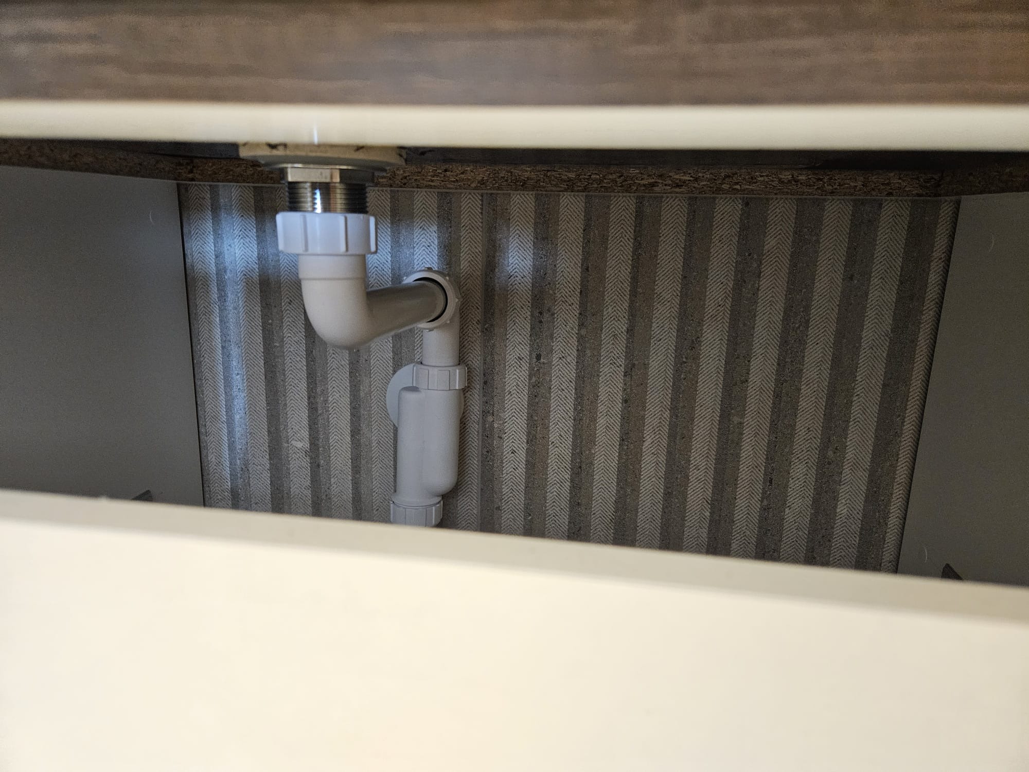 Syfon umywalkowy z białego tworzywa, zamontowany pod blatem szafki, na tle ściany wyłożonej tapetą w szaro-białe pasy. Widok z dołu.