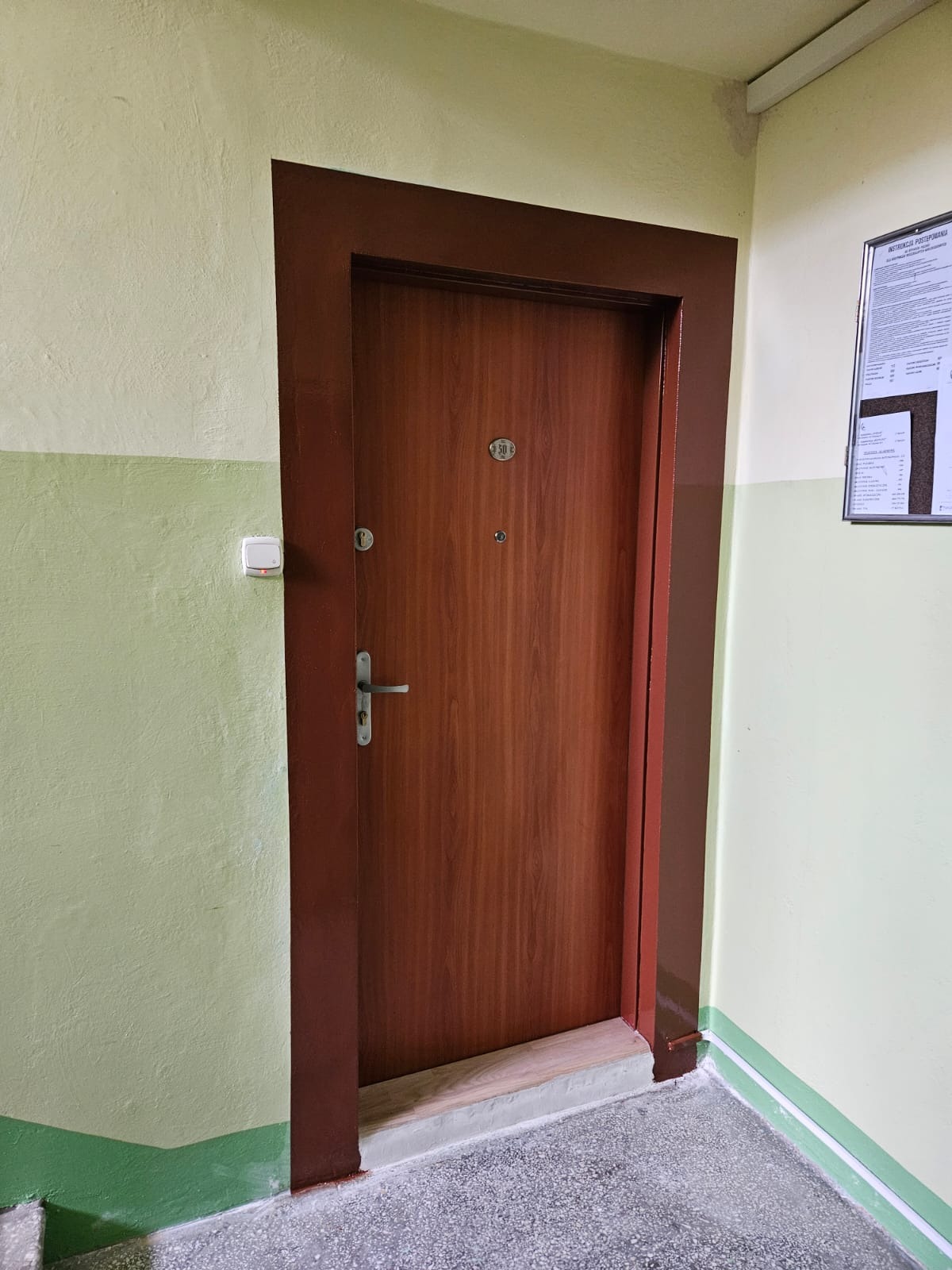 Brązowe, drewnopodobne drzwi wejściowe z numerem '50' w brązowej futrynie, na tle ściany w odcieniach zieleni i beżu, w korytarzu z szarym lastriko.