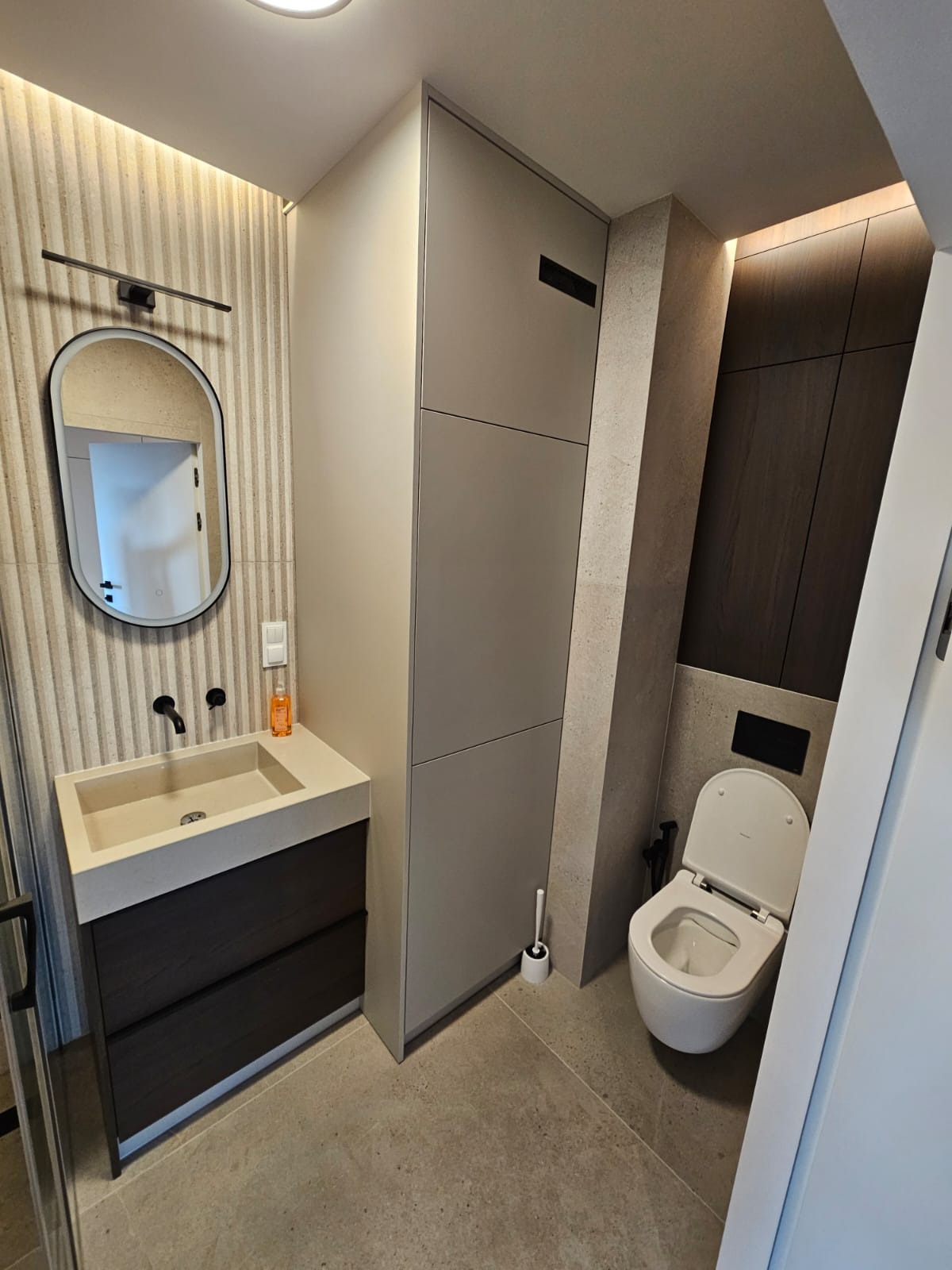 Nowoczesna łazienka z umywalką nablatową, podświetlanym lustrem i zabudową meblową w odcieniach szarości i brązu. Uwagę zwraca oświetlenie LED i minimalistyczny design.