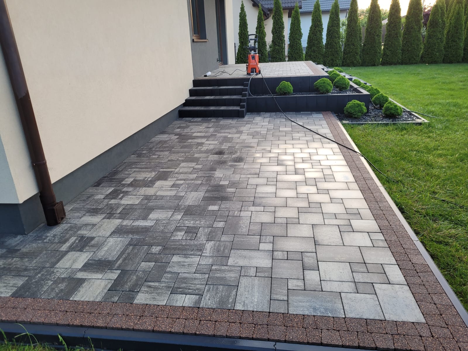 Ułożona kostka brukowa w odcieniach szarości z brązowym obramowaniem, przylegająca do budynku z zielonym trawnikiem w tle. Widoczna myjka ciśnieniowa.