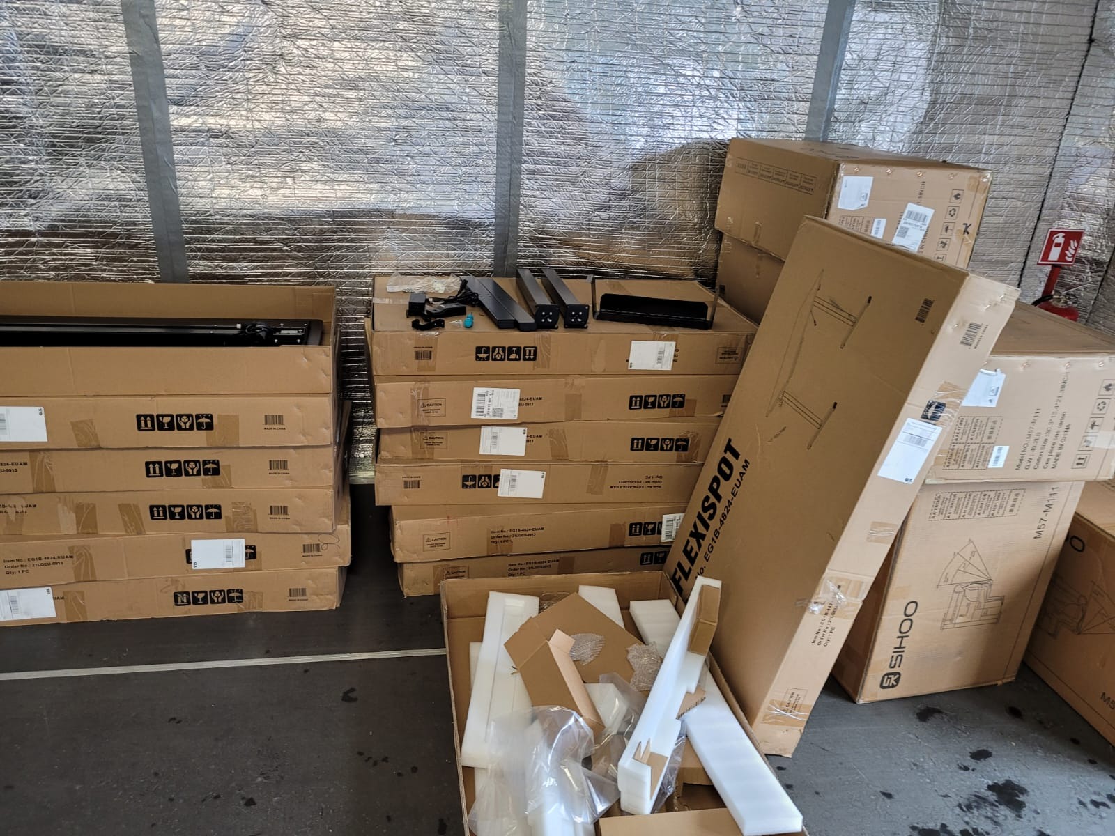 Stosy kartonowych pudeł z elementami biurka Flexispot i Sihoo, gotowych do montażu, na tle ściany z folii aluminiowej. Widoczne akcesoria i instrukcje.