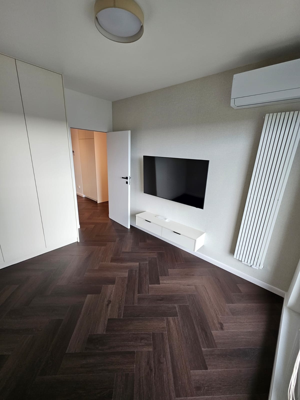 Nowoczesne wnętrze: parkiet w jodełkę, biała szafa wnękowa, minimalistyczna szafka RTV pod wiszącym telewizorem, grzejnik panelowy i klimatyzacja.