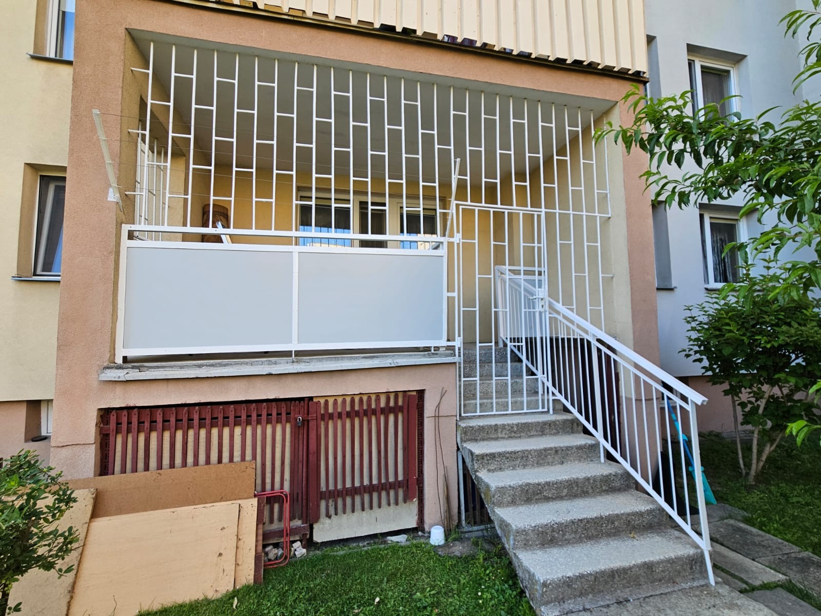 Balkon z białą, nowoczesną balustradą i kratą, schody wejściowe. Widoczne elementy elewacji budynku i zieleń. Ujęcie z dołu, podkreślające geometrię konstrukcji.