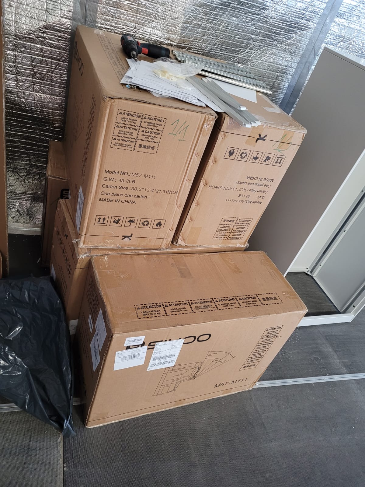 Stos kartonów z modelem M57-M111, wiertarka akumulatorowa i metalowe profile leżące na wierzchu. Przygotowanie do montażu w pomieszczeniu z izolacją.