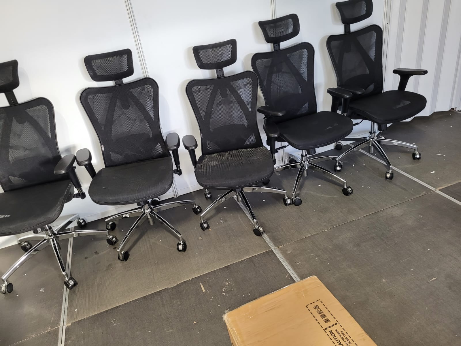 Rząd czarnych, siatkowych krzeseł biurowych z zagłówkami i chromowanymi podstawami na kółkach, ustawionych na szarej podłodze, z kartonem w rogu.
