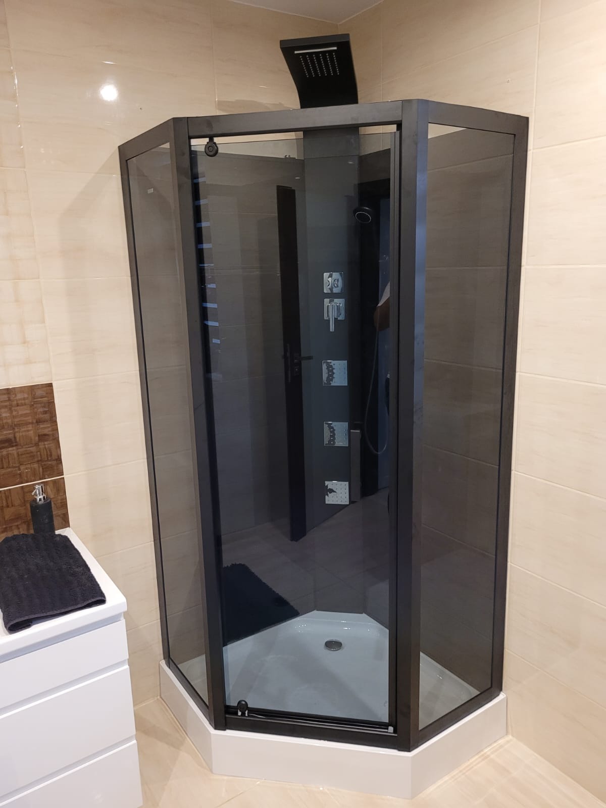 Nowoczesna kabina prysznicowa z czarnymi profilami i deszczownicą w wykończonej łazience z beżowymi płytkami i białą szafką umywalkową.