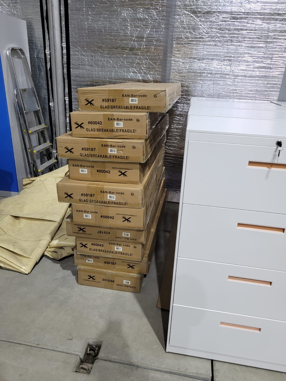 Stos kartonów z oznaczeniami 'GLAS!BREAKABLE!FRAGILE!' obok białej komody z szufladami w surowym wnętrzu. Widoczna drabina i folia izolacyjna na ścianie.