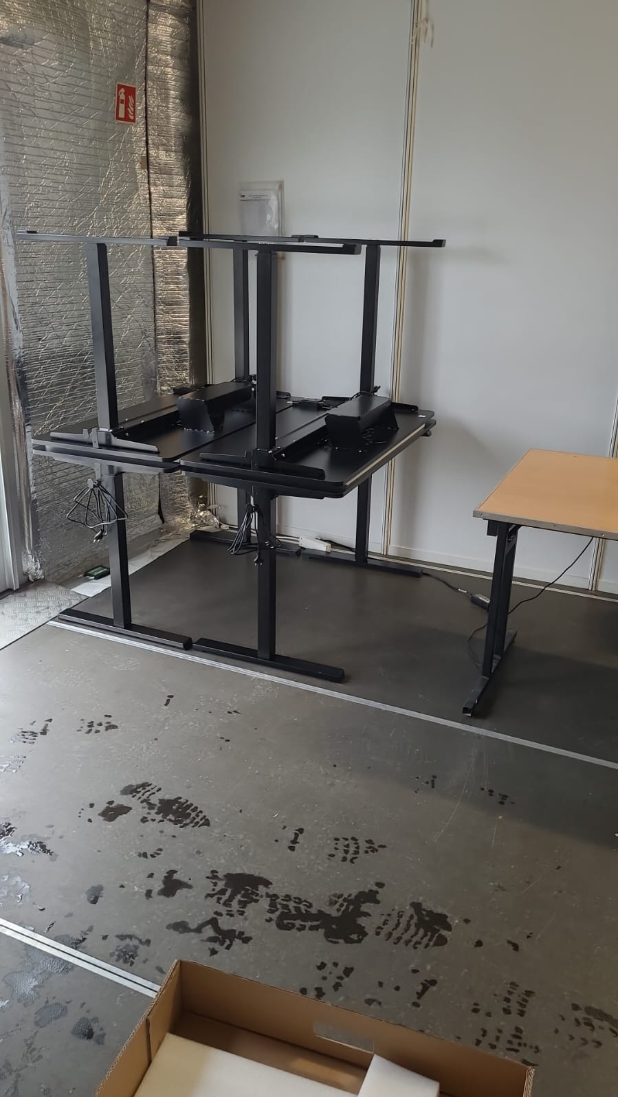 Montaż czarnego, regulowanego biurka z metalową ramą. Widoczne kable i elementy konstrukcyjne. Podłoga z plamami i śladami. Częściowo zmontowany mebel.