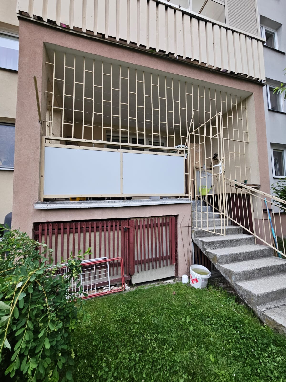 Zabezpieczony balkon z metalową kratą i białymi panelami. Widoczne schody wejściowe i fragment zielonego trawnika. Budynek mieszkalny w tle.