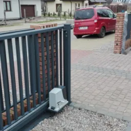 Szara, przesuwna brama wjazdowa z automatyką, zamontowana na ceglanym słupku, widoczny mechanizm napędowy, kostka brukowa na podjeździe, czerwony samochód w tle.