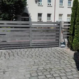 Automatyczna brama przesuwna z poziomych desek w odcieniach szarości, osadzona na kamiennych słupkach, z widocznym napędem, na podjeździe z kostki brukowej, z czerwonym samochodem dostawczym w tle.