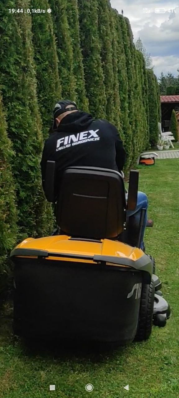 Operator na żółto-czarnej kosiarce siedzącej kosi trawę przy wysokim, gęstym, przyciętym żywopłocie; na plecach bluzy logo firmy ogrodniczej.