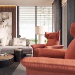 Cooper Estate Sp. z o .o. - Jasne, nowoczesne wnętrze salonu z szarą sofą, okrągłym stolikiem kawowym z książkami i dwoma pomarańczowymi fotelami typu uszak na pierwszym planie, widok na zieleń za oknem z żaluzjami.