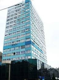 Błękitny wieżowiec z widocznym 14. piętrem, częściowo przysłonięty przez niższą, ciemną konstrukcję z przeszkloną fasadą.