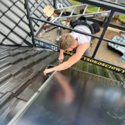 Instalator na dachu z ciemnej dach&oacute;wki, montuje czarny panel słoneczny, widoczny z g&oacute;ry z platformy roboczej.