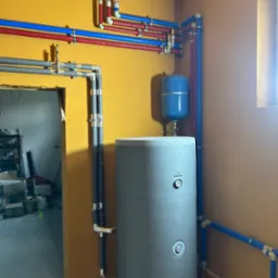 Szary bojler z niebieskim naczyniem wzbiorczym i instalacją wodną z rur czerwonych i niebieskich na żółtej ścianie, widok na surowy stan pomieszczenia.