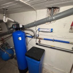 AquaSpark Paweł Kowalczyk - Niebieski filtr wody z czarnym panelem sterowania i zbiornik na sól w piwnicy, widoczne rury instalacji wodnej i fragment instalacji elektrycznej.