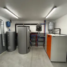 Kotłownia z instalacją grzewczą i zbiornikami w Ostrowsku. Widoczne rury, zawory i osprzęt. Nowoczesny piec CO z pomarańczowym akcentem. Szare ściany i betonowa posadzka.