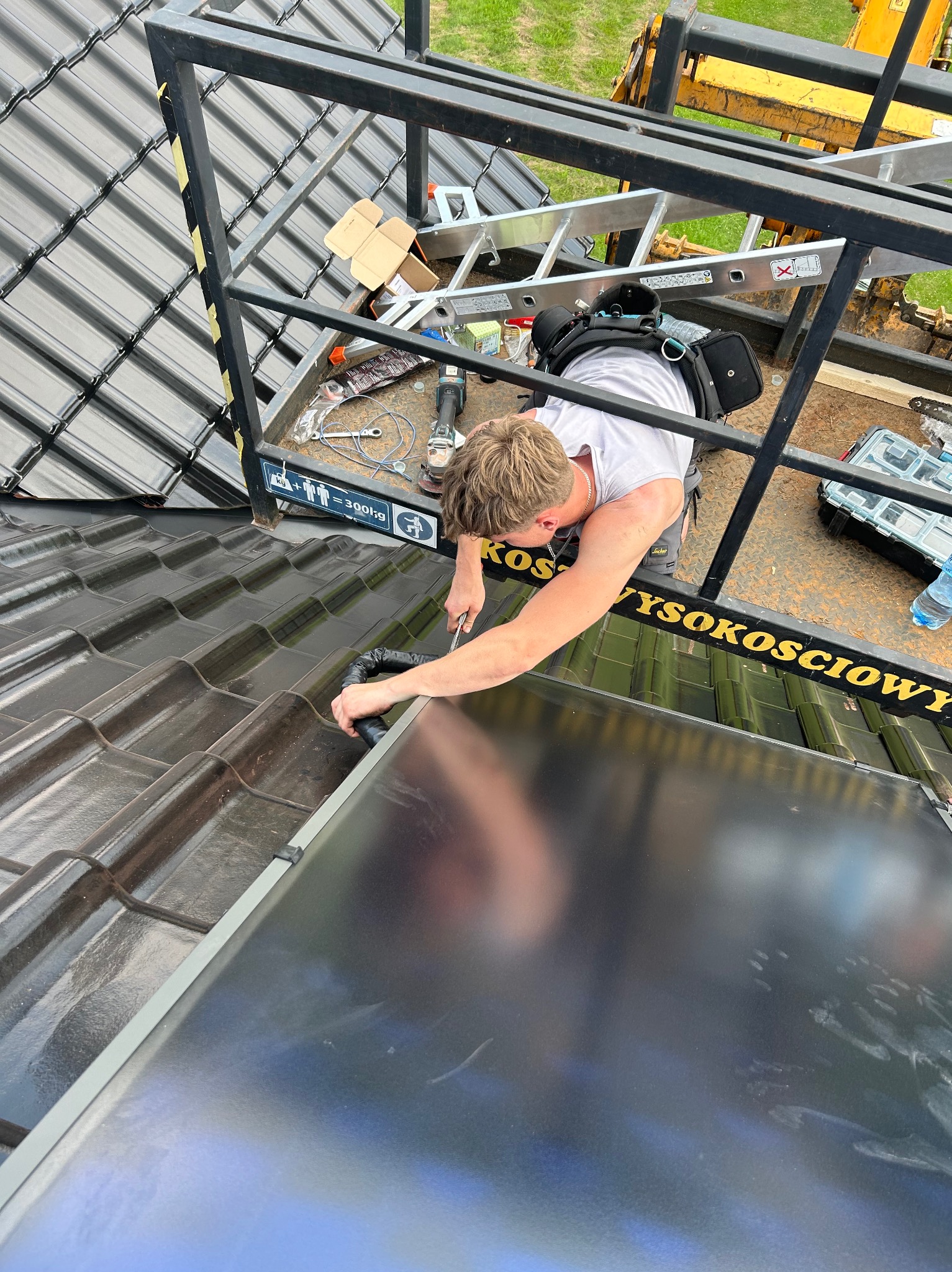 Instalator na dachu z ciemnej dachówki, montuje czarny panel słoneczny, widoczny z góry z platformy roboczej.