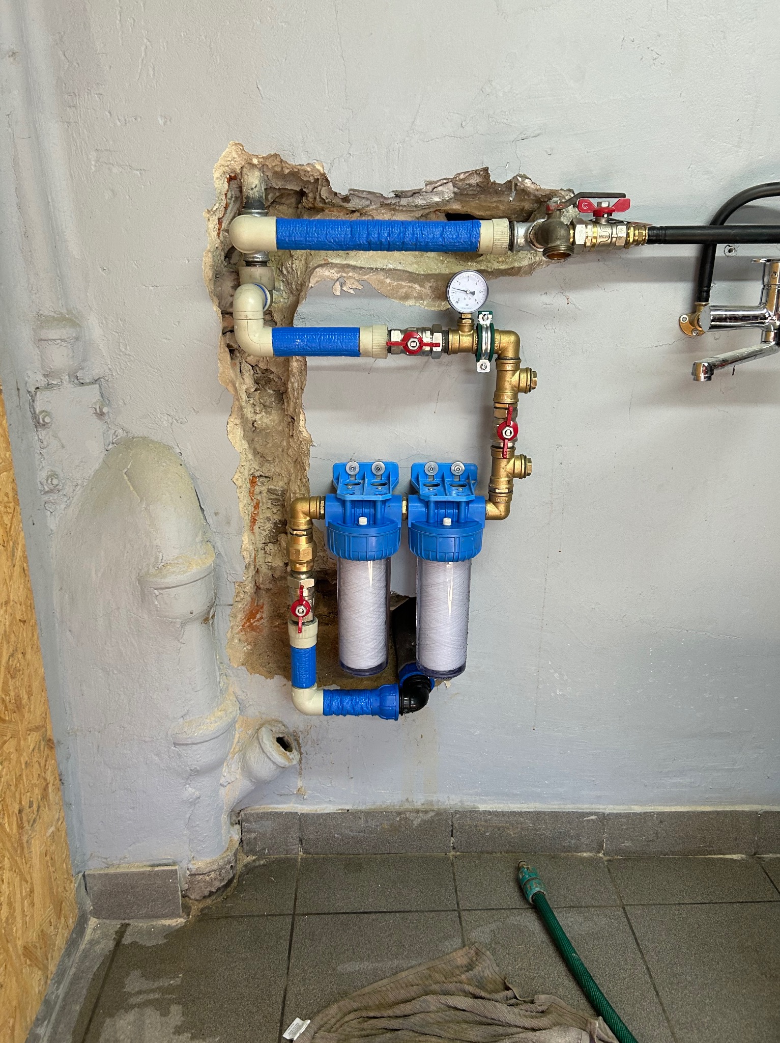 Dwa filtry wody zamontowane na ścianie z widocznymi rurami, zaworami i manometrem. Ściana w stanie surowym z widocznymi uszkodzeniami tynku.