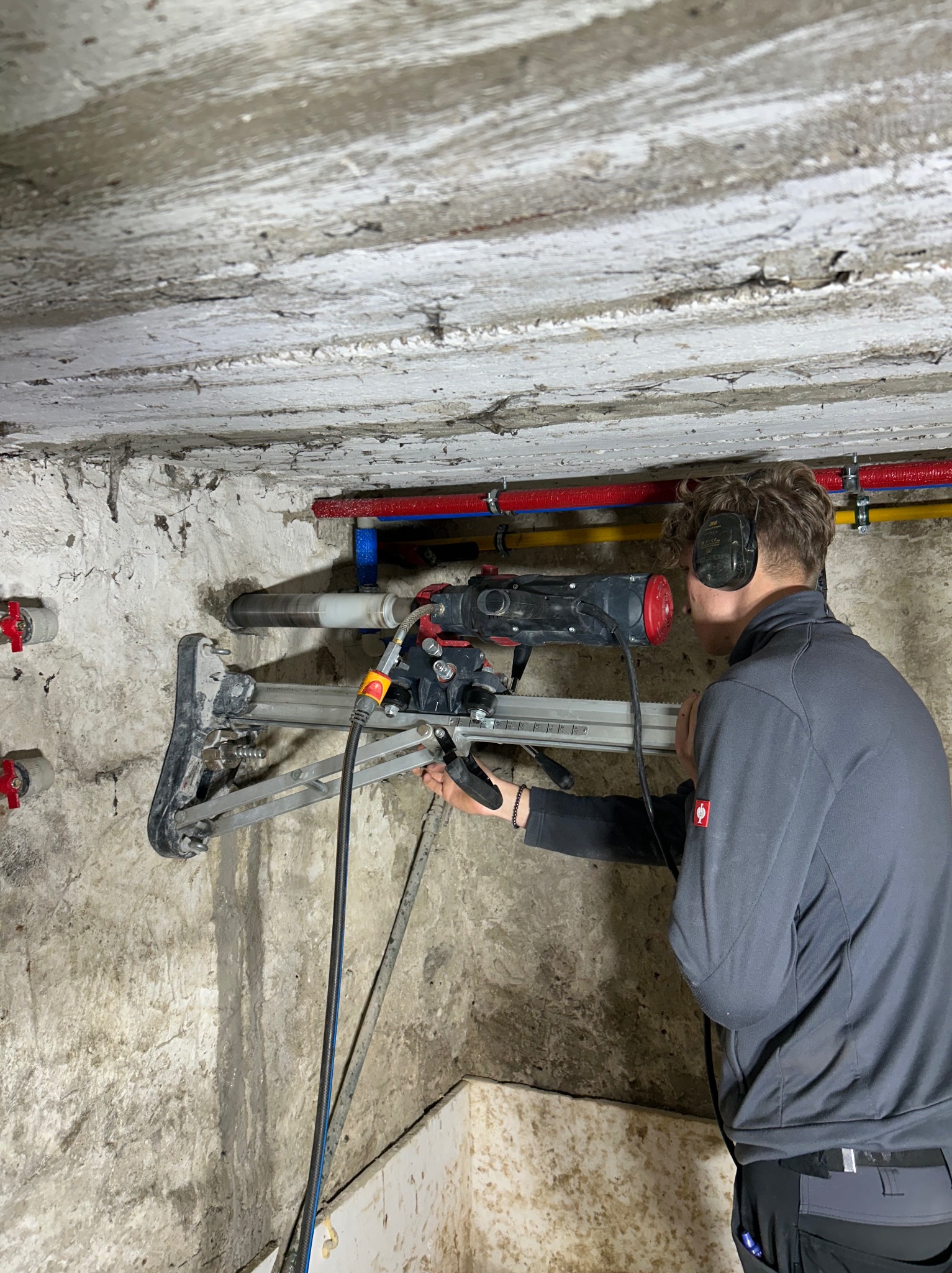 Pracownik w słuchawkach wierci otwór w ścianie w Ostrowsku za pomocą wiertnicy diamentowej, widoczne rury instalacji wodnej.