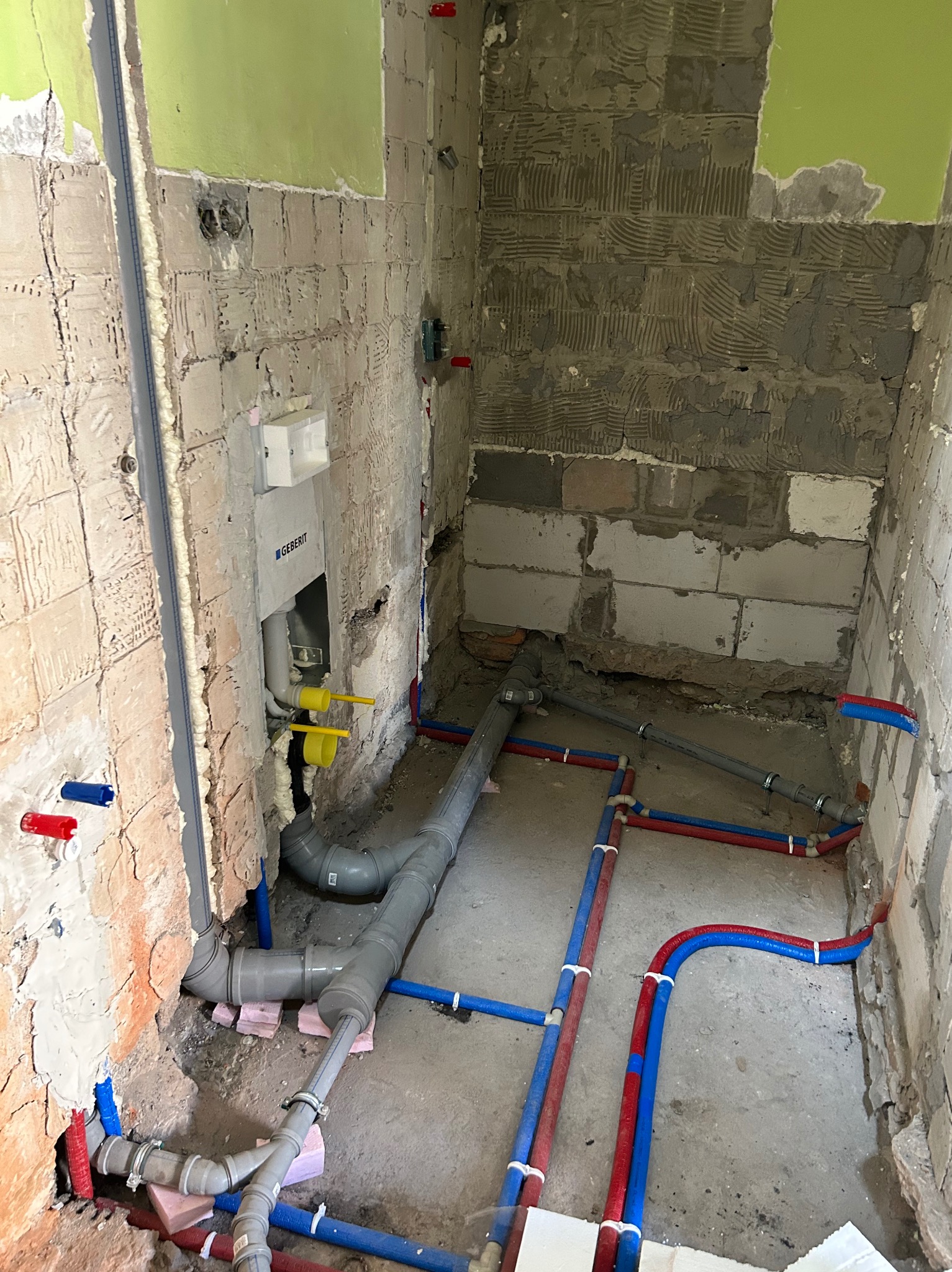 Surowy stan łazienki w trakcie instalacji hydraulicznej: widoczne rury kanalizacyjne z szarego PCV, rury doprowadzające wodę w kolorach czerwonym i niebieskim, zamontowany stelaż podtynkowy WC...
