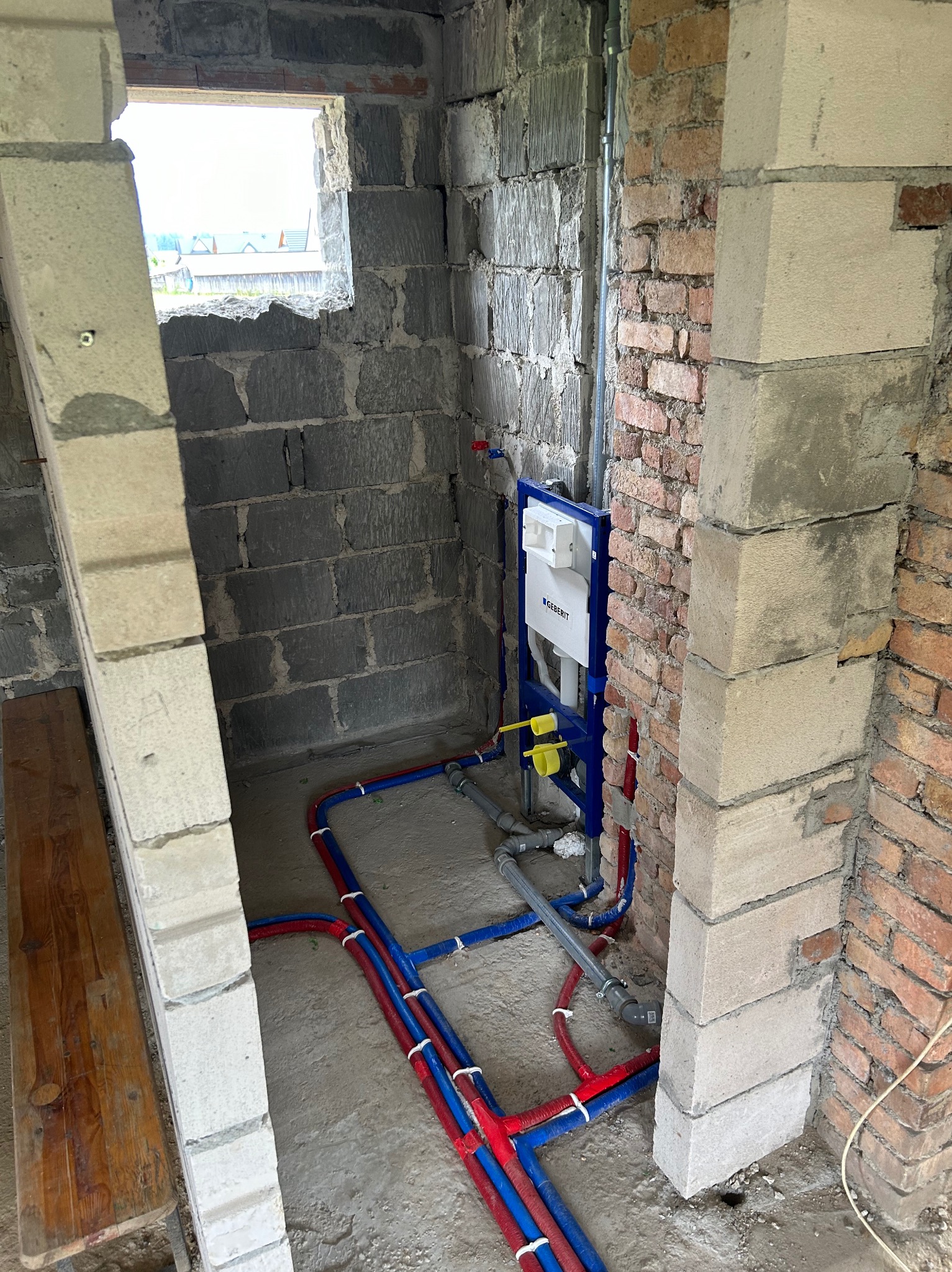 Surowy stan łazienki w trakcie remontu: widoczne instalacje wodno-kanalizacyjne z niebieskimi i czerwonymi rurami, stelaż podtynkowy Geberit zamontowany na ceglanej ścianie, surowe ściany z betonu...