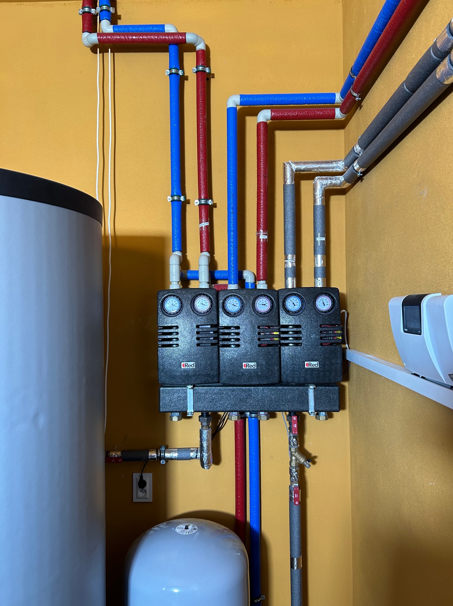 Rozdzielacz ogrzewania podłogowego z rurami w kolorach niebieskim i czerwonym, manometry wskazują ciśnienie, zbiornik wyrównawczy i bojler w tle, żółta ściana.