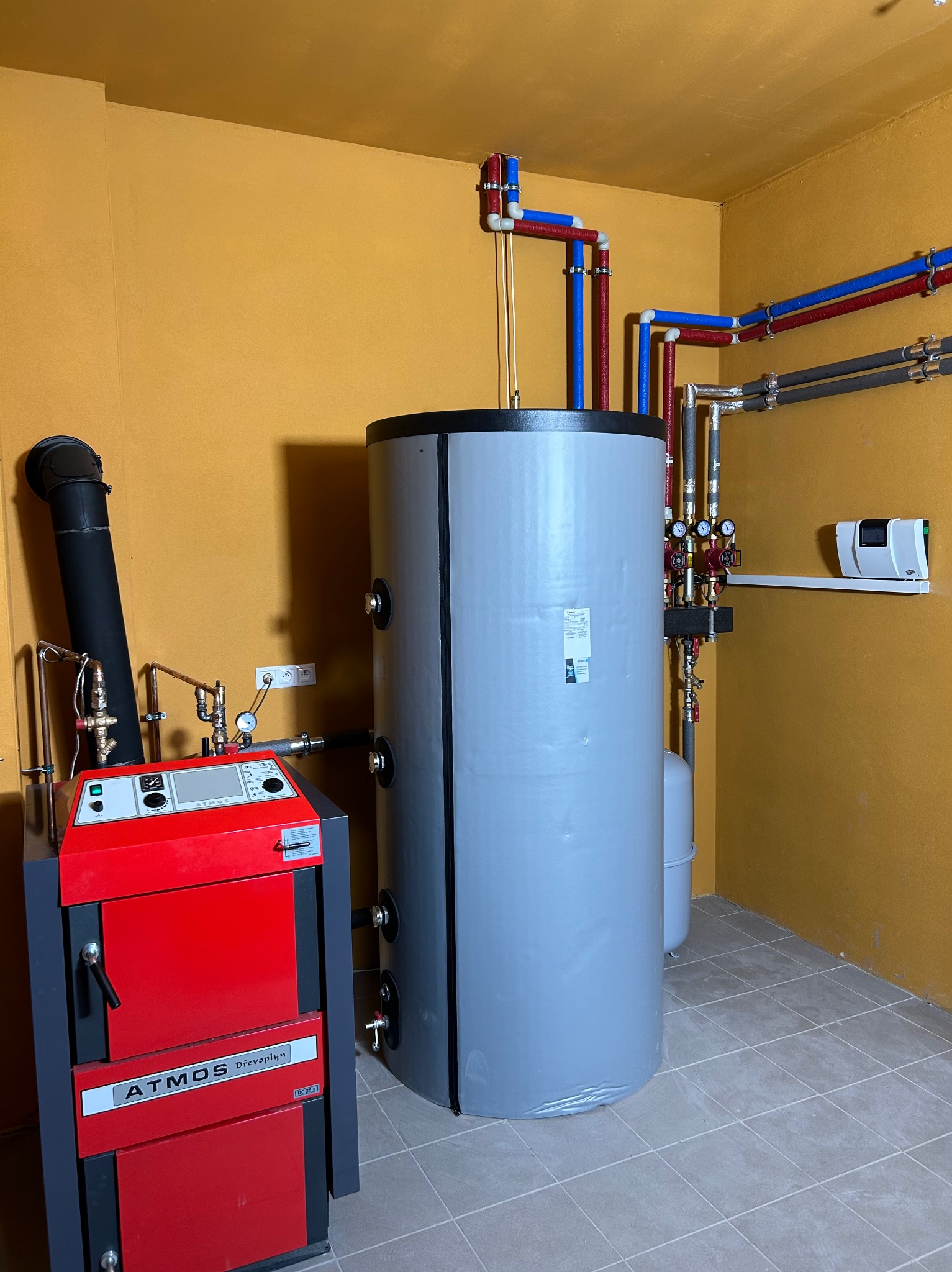 Kotłownia z piecem Atmos i szarym zbiornikiem akumulacyjnym, widoczne rury instalacji grzewczej w kolorach czerwonym i niebieskim, pomieszczenie wykończone w kolorze pomarańczowym, szare płytki...