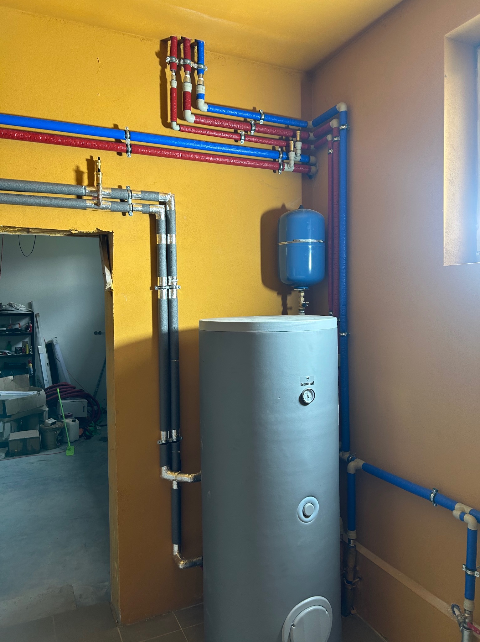Szary bojler z niebieskim naczyniem wzbiorczym i instalacją wodną z rur czerwonych i niebieskich na żółtej ścianie, widok na surowy stan pomieszczenia.