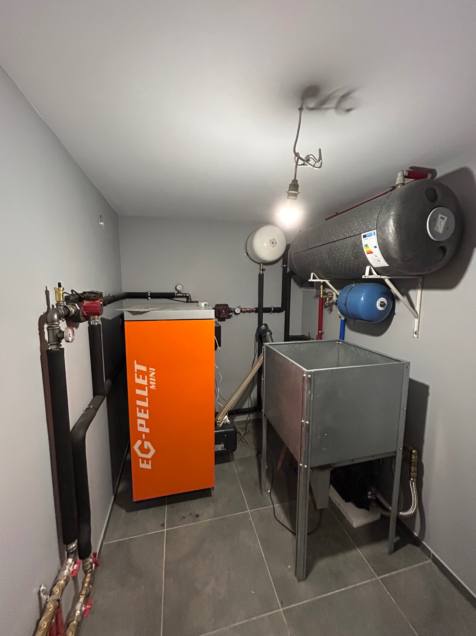 Pomieszczenie kotłowni z piecem na pellet EG-Pellet Mini, zbiornikiem buforowym, naczyniem wzbiorczym i instalacją hydrauliczną, oświetlone pojedynczą żarówką.
